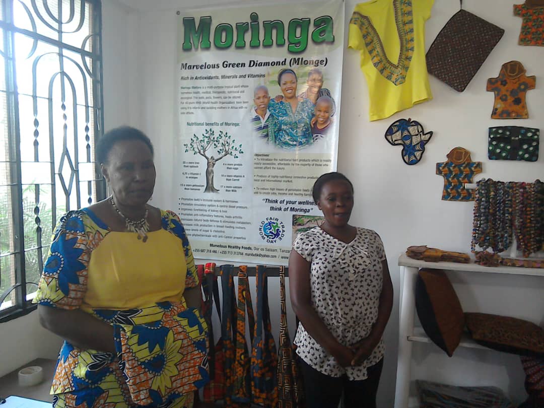 Two women standing beside a Moringa nutrition display and handmade textile goods.